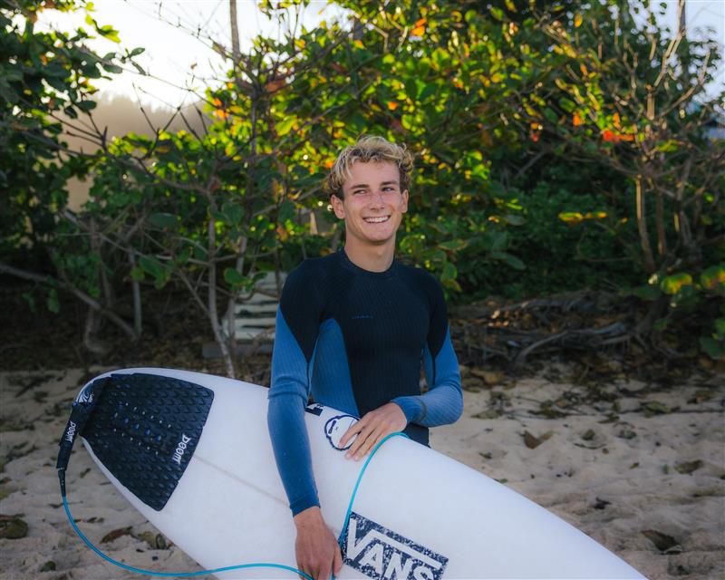 Person holding a surfboard with a wetsuit on, standing on sandy ground with greenery in the background