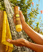Hand holding a brown and yellow bottle labeled 'Luna Sun' under a shower with greenery in the background.