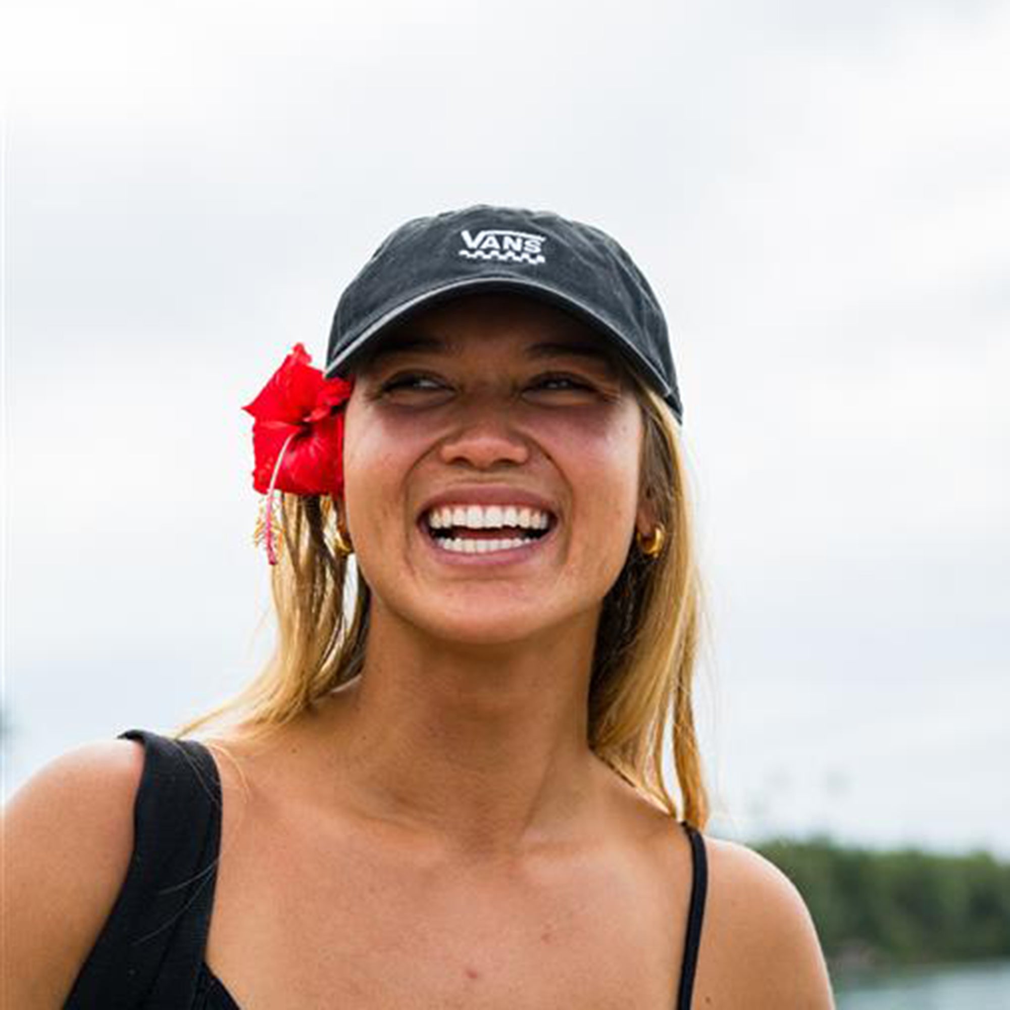 Woman wearing a black Vans cap with a red flower in her hair, smiling outdoors.