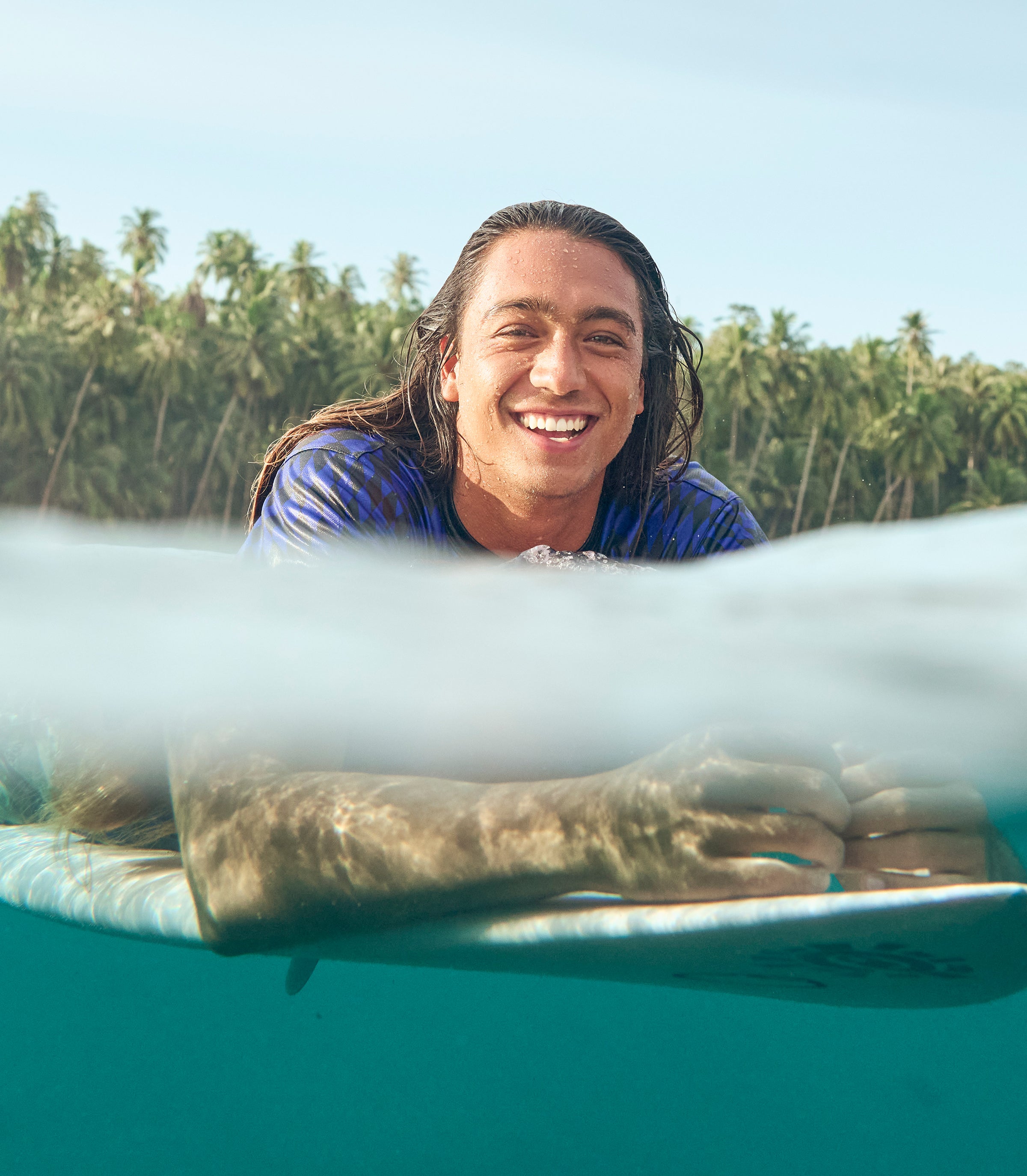 Person lying on a surfboard with a scenic background of trees and clear sky.